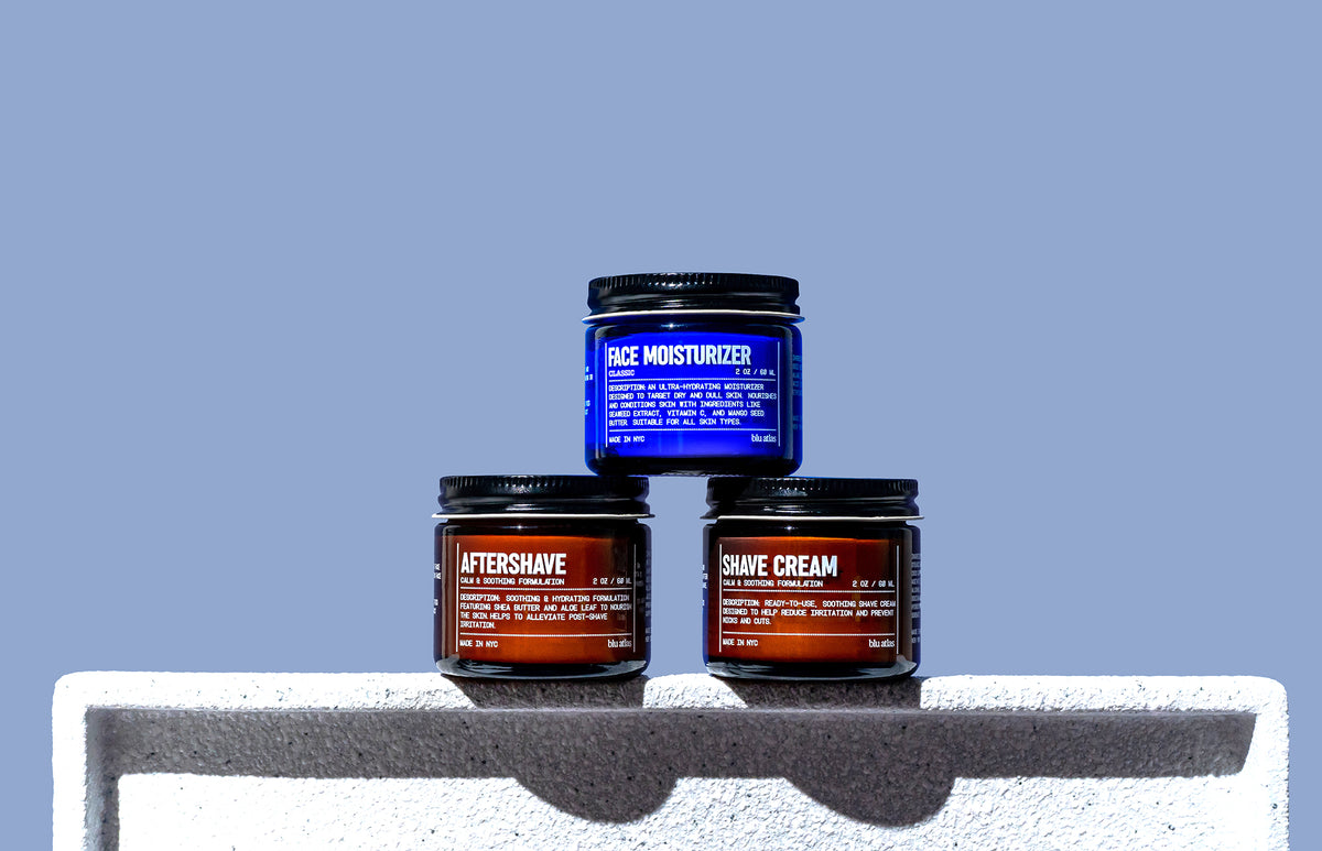 The Blu Atlas Shave Bundle, consisting of jars of face moisturizer, aftershave balm, and shave cream, is elegantly arranged on a white surface with a calming blue background.