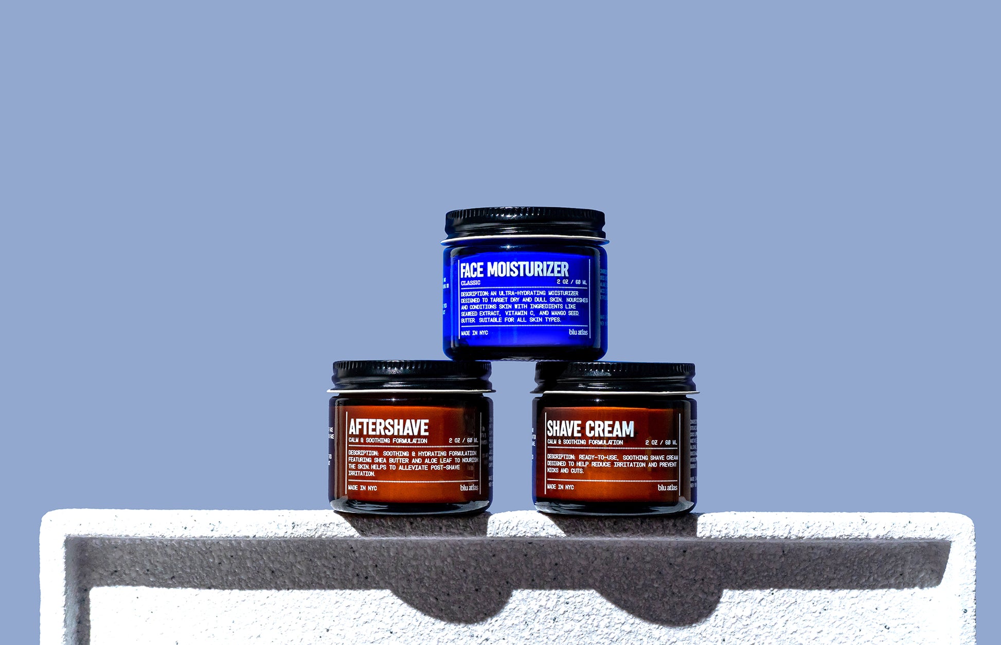 The Blu Atlas Shave Bundle, consisting of jars of face moisturizer, aftershave balm, and shave cream, is elegantly arranged on a white surface with a calming blue background.