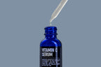 A dropper dispenses liquid into a blue bottle labeled Blu Atlas Vitamin C Serum, celebrated for its powerful antioxidants, against a gray background.