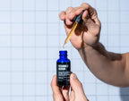 A person holds a dropper above a blue bottle labeled Blu Atlas Vitamin C Serum, a popular antioxidant known for its skin-brightening properties, with a tiled wall in the background.
