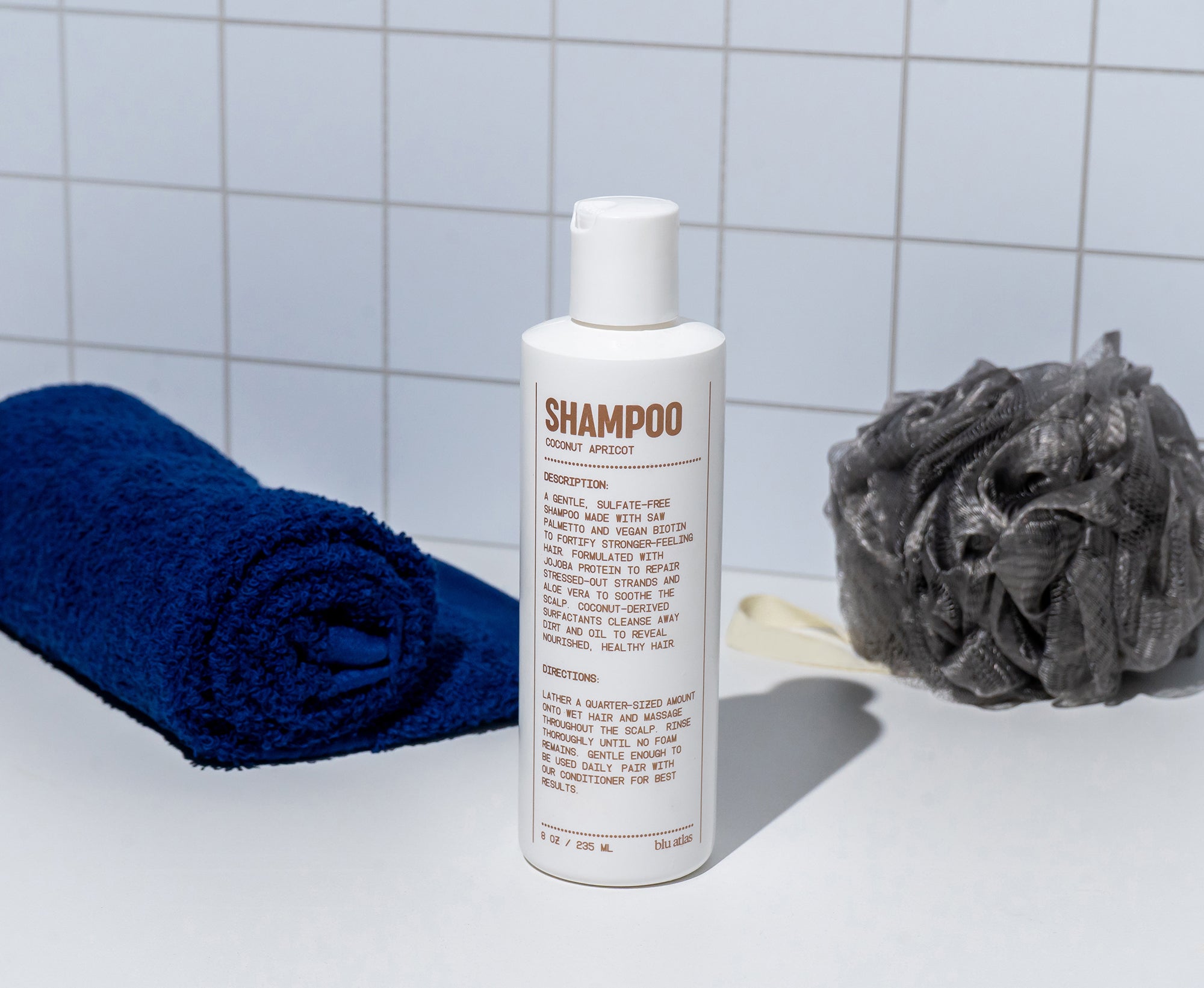 A white bottle of Blu Atlas Shampoo is placed on a surface beside a rolled blue towel and a gray bath pouf, set against a white tiled wall.