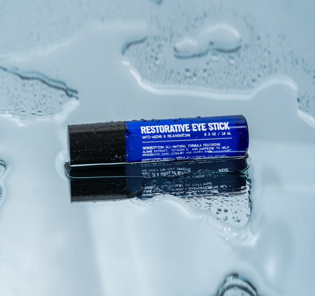 The Blu Atlas Men's Restorative Eye Stick For Puffy Eyes, featuring a black cap and enriched with algae extract, rests on a wet reflective surface.
