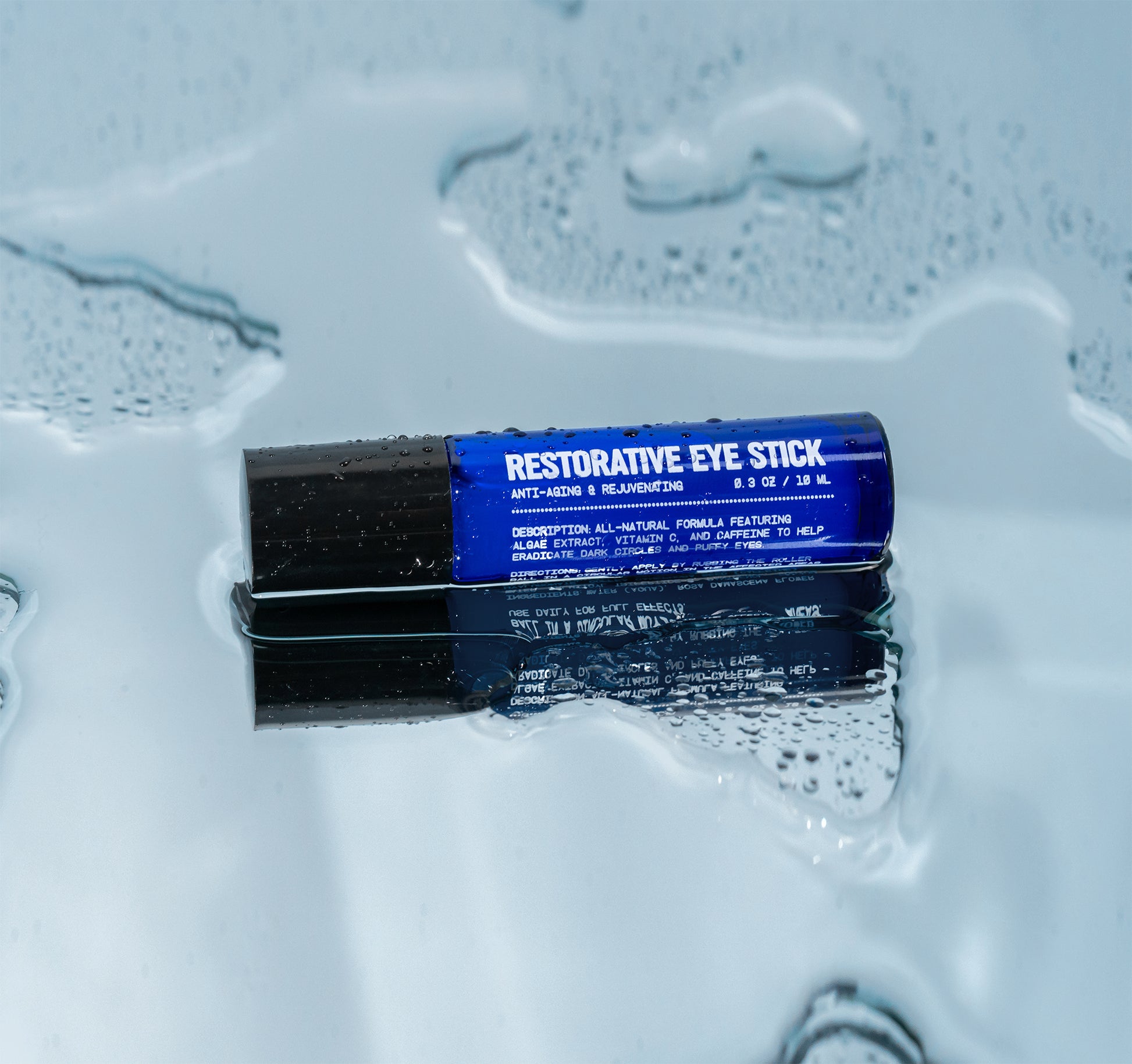 The Blu Atlas Men's Restorative Eye Stick For Puffy Eyes, featuring a black cap and enriched with algae extract, rests on a wet reflective surface.