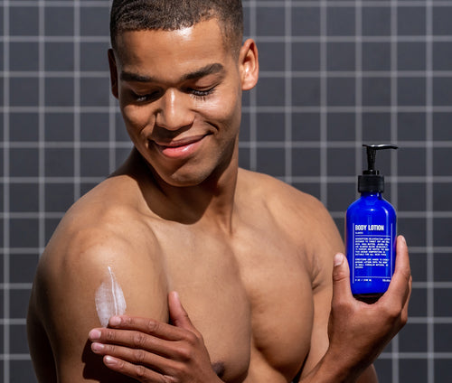 A man applies Blu Atlas Body Lotion to his bare shoulder while holding a blue bottle labeled Body Lotion, standing in front of a tiled wall.