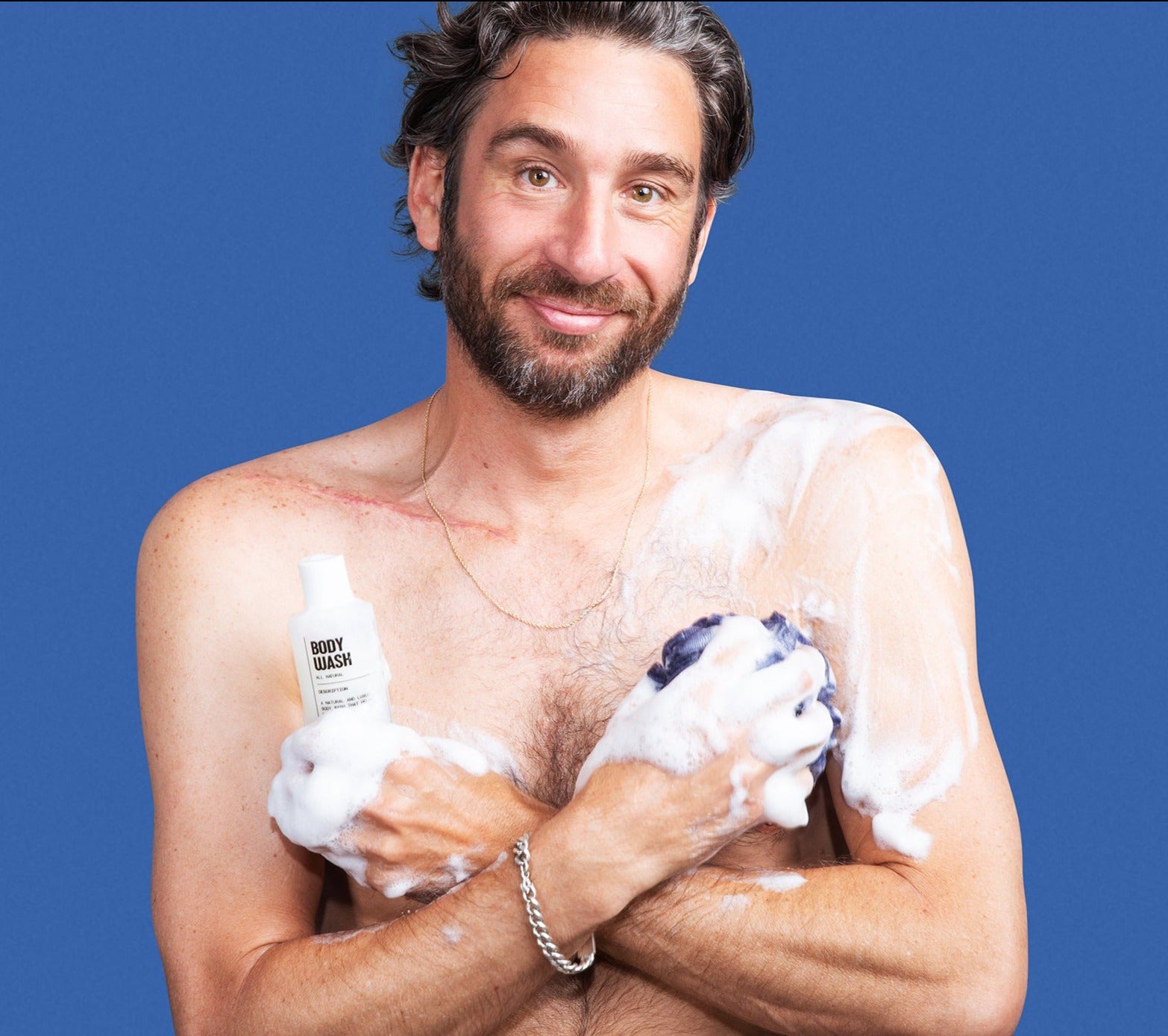 A man uses Blu Atlas Hydrating Body Wash, lathering rich foam on his chest against a blue background.