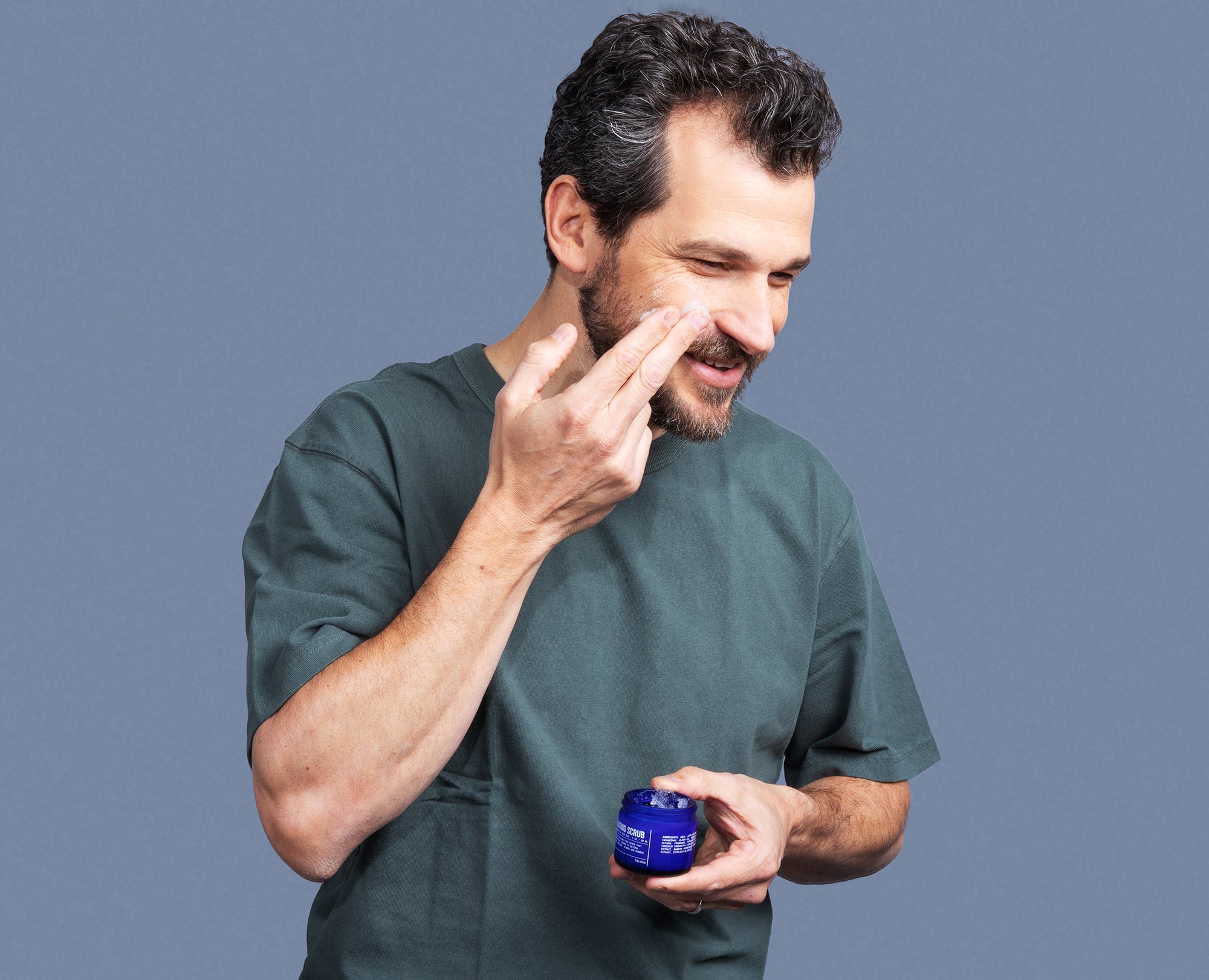 A man wearing a green t-shirt adds Blu Atlass Gentle Exfoliating Scrub for All Skin Types to his grooming routine, applying it with his fingers while holding the small blue container.